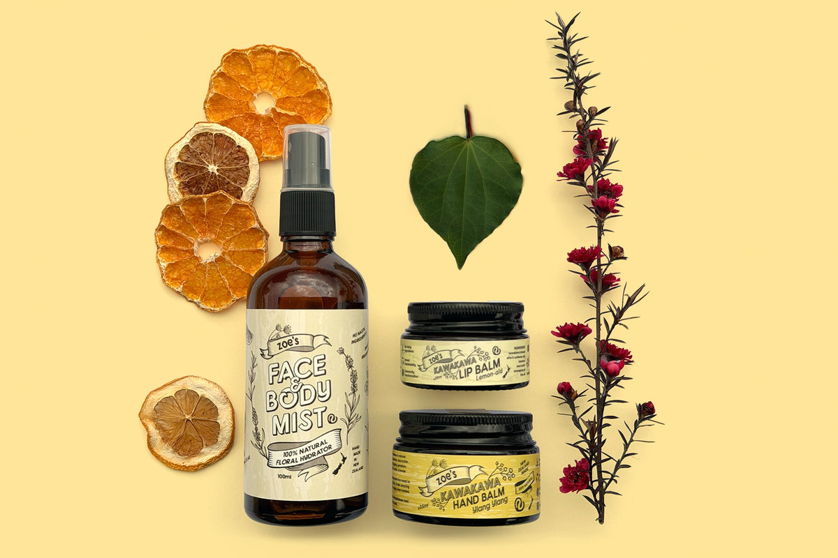 Zoe's Kawakawa Paper Gift Box, A flat-lay product arrangement on a pale yellow background featuring a brown glass spray bottle labeled Zoe's Face Body Mist, two small jars with black lids labelled 'Zoe's Kawakawa Lemon-Aid Lip Balm' and 'Zoe's Kawakawa Ylang Ylang Hand Balm', dried orange slices, a green kawakawa leaf, and a red flowering manuka stem.