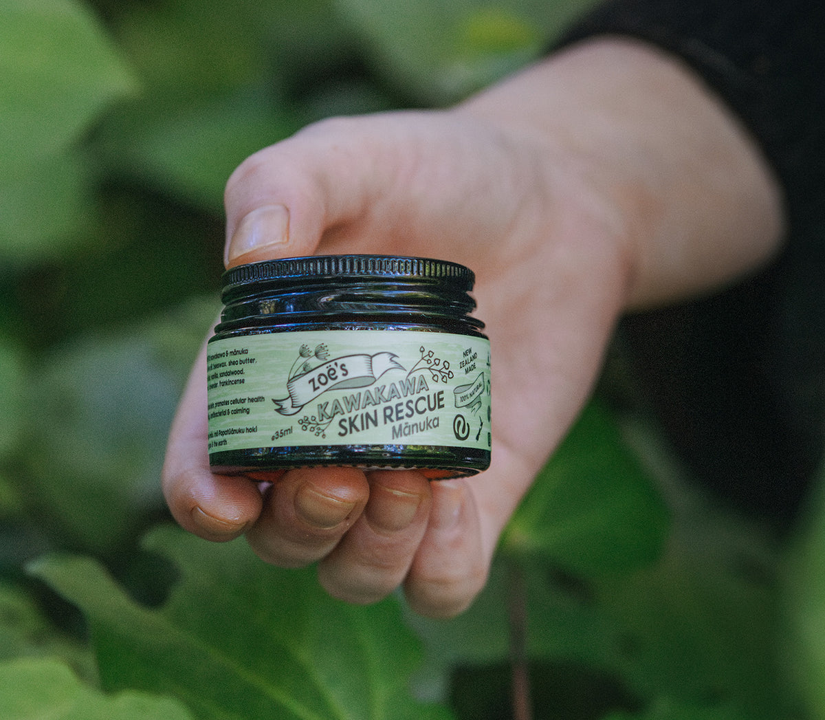 Hand holding a 60ml jar of 'Zoe's Kawakawa Skin Rescue' product against a green leafy kawakawa background